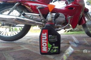 En primer plano una botella de aceite, detrás una moto de 110 cc roja