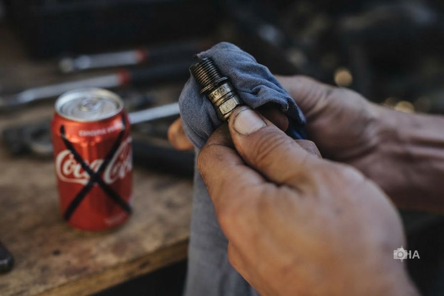 Un mecánico limpia una bujía con un trapo. en segund plano, un latita de Coca Cola tachada con un cruz.