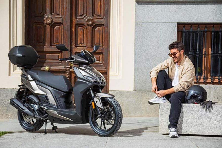 Un joven, sentado frente a unsa casa, ata sus cordones al lado de un scooter Kymco Agility S 125
