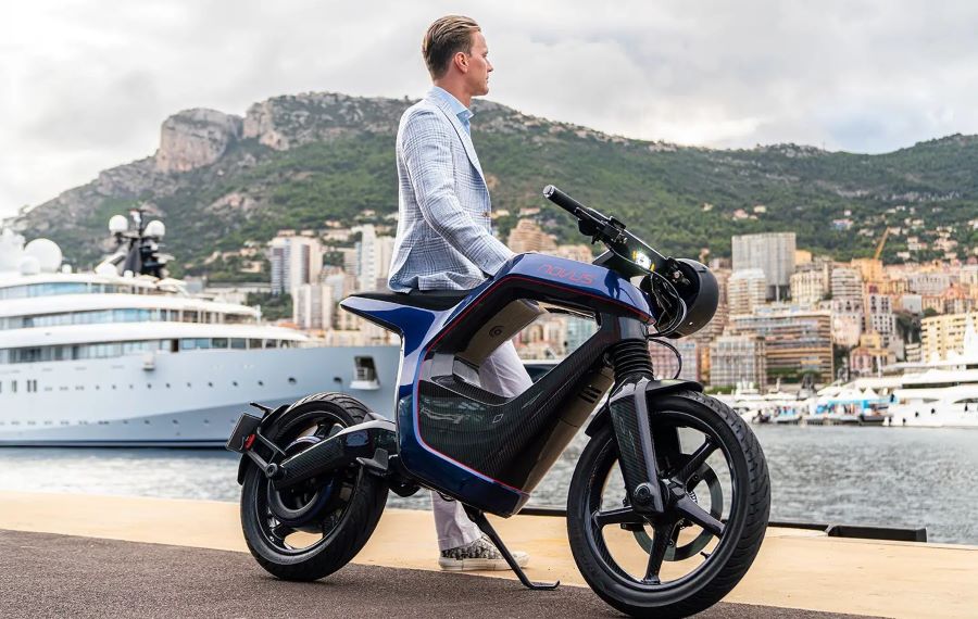 Novus One: carbono, 25 kW y una idea difícil de encasillar Un modelo, con saco celeste claro, posa al lado de una moto eléctrica Novus One sobre un fondode embarcaciones