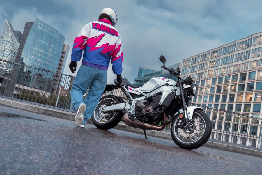 Un piloto con indumentaria al tonos rosa, azul y blanco se acerca a una Yamaha XSR900 2027